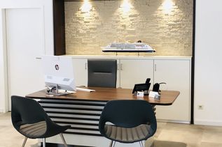 Moderner Arbeitsplatz in einem Reisebüro mit Holztisch, zwei Besucherstühlen und einem Modellkreuzfahrtschiff als Dekoration vor beleuchteter Steinwand.