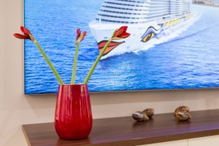 Rote Vase mit drei roten Blüten auf einer Holzanrichte, im Hintergrund ein Bildschirm mit dem Bild eines Kreuzfahrtschiffs auf dem Meer.