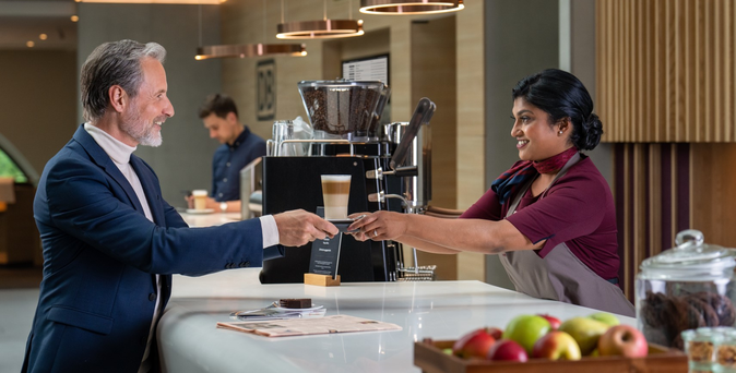 Eine Mitarbeiterin reicht einem Mann im Anzug am Tresen einen Latte Macchiato, im Vordergrund Obstkiste, im Hintergrund eine Kaffeemaschine.