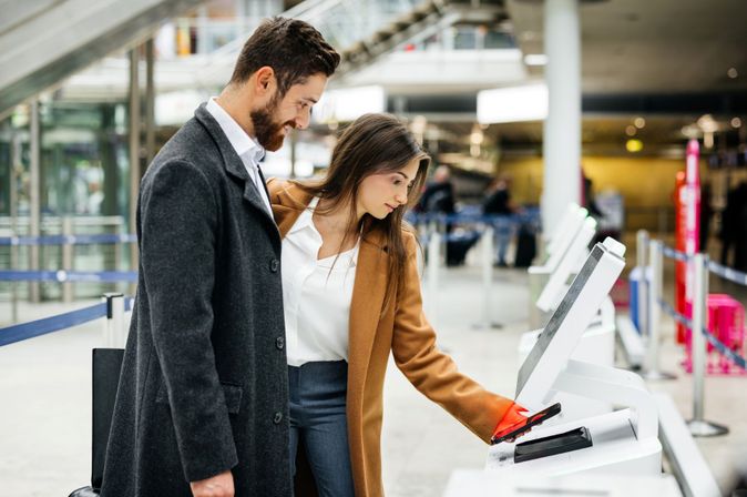 Mann und Frau am Self-Check-in-Automaten im Flughafenterminal; die Frau hält ihr Smartphone an den Scanner