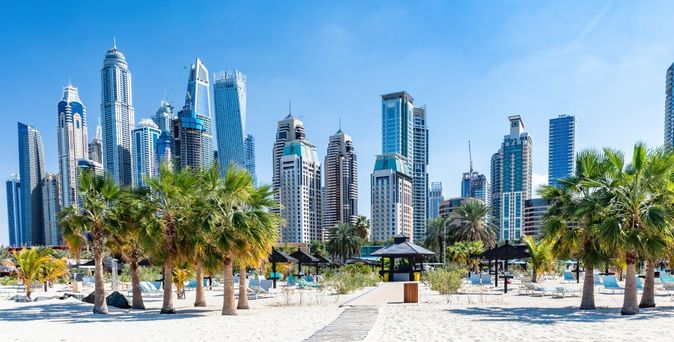 Weißer Sandstrand mit Palmen und Sonnenliegen vor der beeindruckenden Skyline von Dubai Marina bei strahlend blauem Himmel.