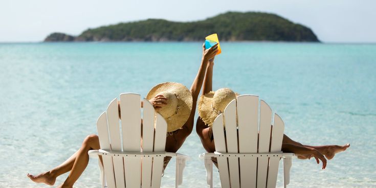 Zwei Personen mit Strohhüten entspannen in weißen Liegestühlen am Strand und stoßen mit bunten Drinks auf das türkisfarbene Meer hinaus an – Sinnbild für Sommer, Leichtigkeit und unbeschwerten Genuss im Urlaub.
