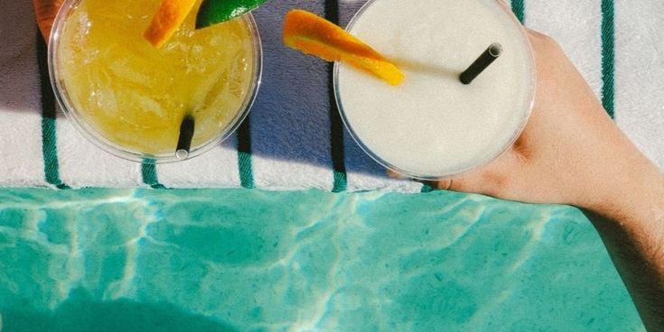 Zwei sommerliche Cocktails mit Zitrusgarnitur stehen auf einem gestreiften Handtuch am türkisblauen Pool.