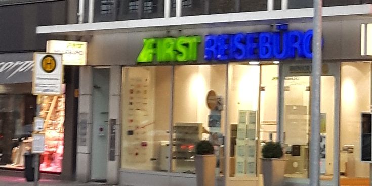 Außenansicht des FIRST Reisebüros an der Bismarckstraße in Mönchengladbach mit Glasfassade und beleuchtetem Schriftzug.