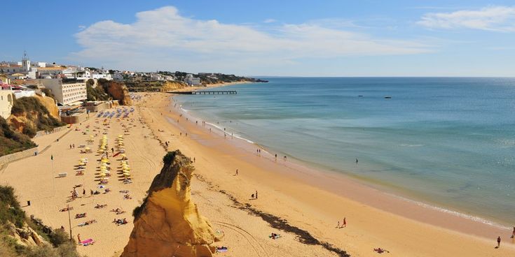 Weiter Sandstrand an der Algarve mit Badegästen, gelben Sonnenschirmen und markanter Felsformation vor ruhigem Meer.