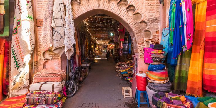 Bunter Basar in Marrakesch mit Teppichen, Stoffen und traditionellen Waren unter einem gemauerten Torbogen.