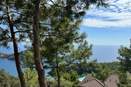 Kiefern vor bewaldetem Hang mit Hausdächern und Blick auf das Meer unter blauem Himmel mit Wolken