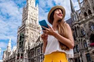 Frau mit Smartphone und Sonnenhut steht vor dem Neuen Rathaus in München und lächelt in den Himmel.