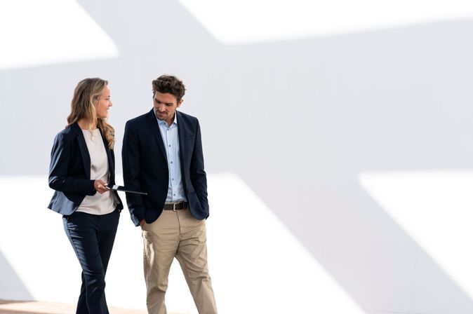 Zwei Geschäftsleute in Businesskleidung gehen nebeneinander und sprechen; die Frau hält ein Tablet in der Hand; vor hellem, minimalistischem Hintergrund.