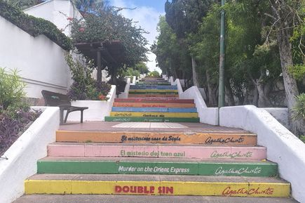 Bunte Außentreppe mit Buchtiteln und Autorennamen auf den Stufen, umgeben von Bäumen und weißen Mauern unter blauem Himmel.