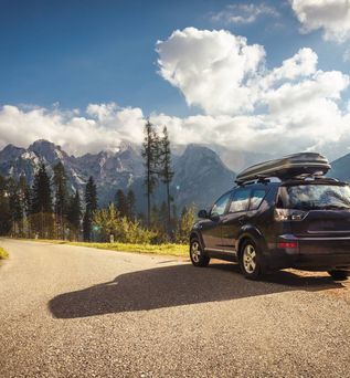 Ein dunkler SUV mit Dachbox steht auf einer kurvigen Bergstraße vor einer Kulisse aus Nadelbäumen und schroffen Alpenbergen unter blauem Himmel mit weißen Wolken.