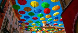 Zahlreiche bunte Regenschirme schweben über einer schmalen Straße zwischen farbenfrohen Häusern.