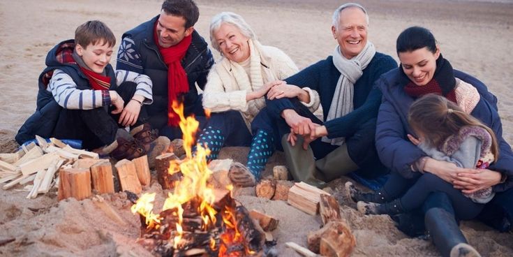 Mehrgenerationenfamilie sitzt lachend am Strand um ein Lagerfeuer – gemeinsame Auszeit mit Wärme, Nähe und Urlaubsfreude.