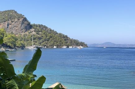 Strand mit Sonnenschirmen aus Stroh, Palmenblättern im Vordergrund, dahinter ruhiges Meer und bewaldeter Hügel unter blauem Himmel.