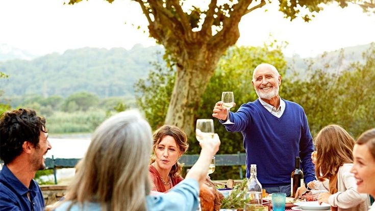 Mehrere Personen sitzen an einem gedeckten Tisch im Freien und stoßen mit einem Glas Wein an