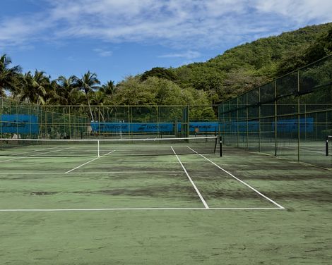 Leerer Tennisplatz in tropischer Umgebung mit Palmen und einem bewaldeten Hügel unter blauem, wolkigem Himmel.