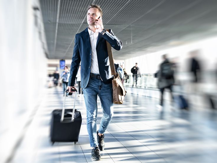 Geschäftsmann mit Trolley und Handy an trubeligem Flughafen