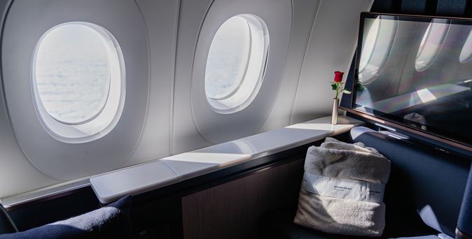 Lufthansa First-Class-Sitz mit zwei Fensterluken, roter Rose auf der Ablage, gefaltetem Handtuch und Bildschirm.