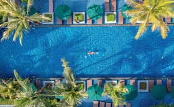 Luftaufnahme eines tropischen Resortpools mit Palmen, grünen Sonnenschirmen und Liegen; eine Person schwimmt im türkisblauen Wasser.