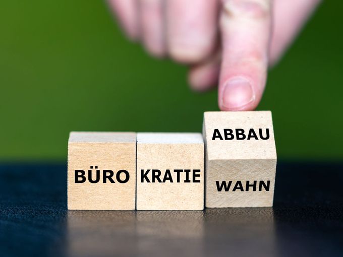 Eine Hand dreht den dritten von drei nebeneinanderstehenden Holzwürfeln um. Die Würfel bilden zusammen entweder das Wort „Bürokratiewahn" oder „Bürokratieabbau" – je nach Stellung des dritten Würfels. Hintergrund ist grün und unscharf.