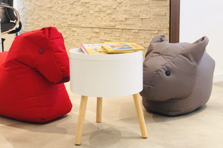 Gemütliche Kinderspielecke im Reisebüro mit roten und grauen Sitzsäcken in Tierform, rundem Tisch und Kinderbüchern.