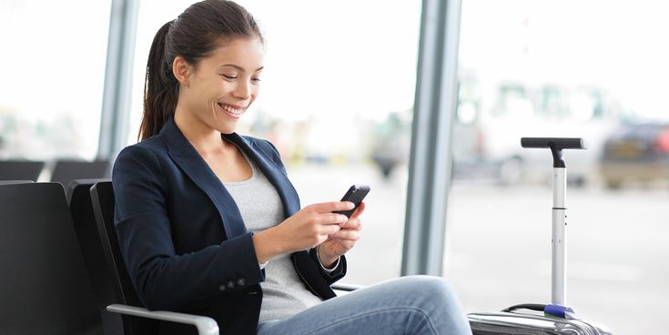 Frau sitzt mit Handy und Trolley am Flughafen