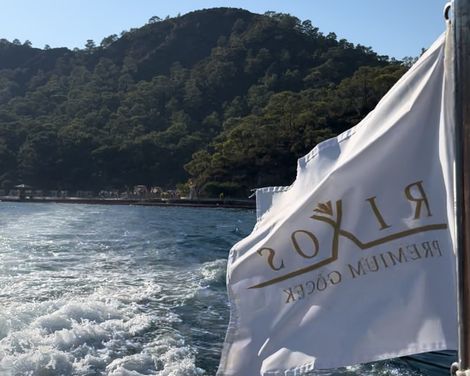 Weißes Flaggenbanner mit goldener Aufschrift 'RIXOS PREMIUM GÖCEK' weht auf einem Boot, im Hintergrund bewaldeter Hügel und Wasser mit Bootsbewegung.