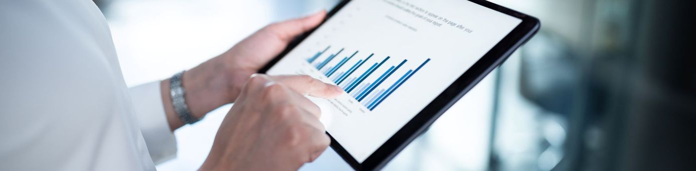 Eine Person in weißem Hemd hält ein Tablet in beiden Händen und tippt mit dem Zeigefinger auf ein Balkendiagramm, das auf dem Display zu sehen ist. Im Hintergrund ein heller, verschwommener Büroflur.