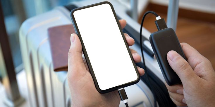 Männerhände halten links ein Smartphone und rechts eine Powerbank. Im Hintergrund ein Koffer in einer Airport Lounge.