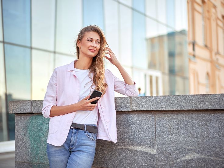 Blonde Frau mit rosa Bluse und Handy lehnt entspannt an Mauer