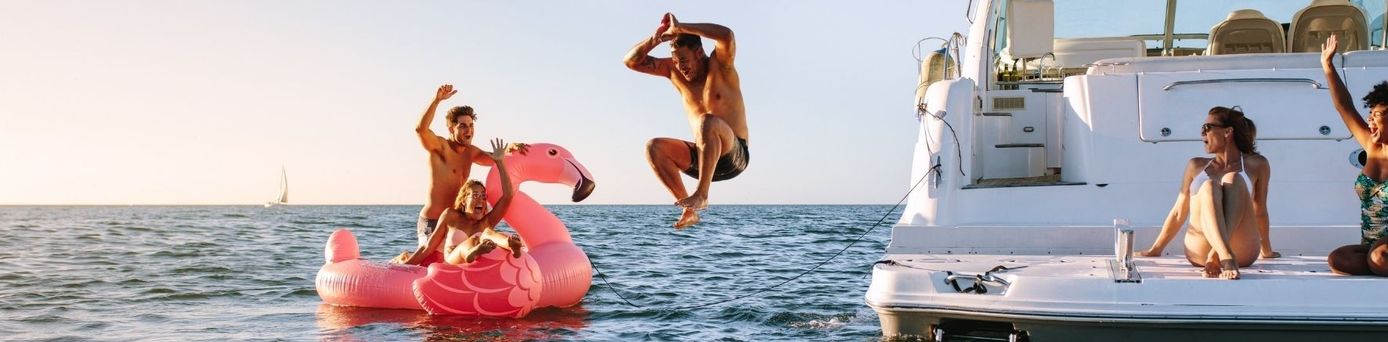 Freunde feiern auf einer Yacht und springen neben einem großen pinken Flamingo ins Meer.