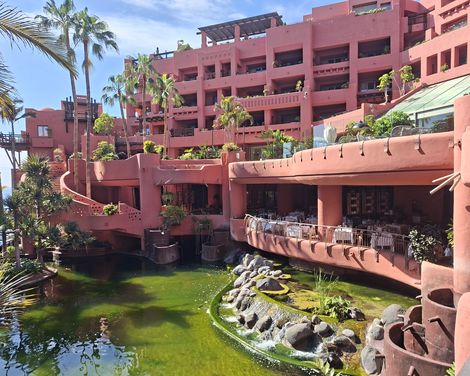 Terrakottafarbene Hotelanlage des Ritz-Carlton mit Balkonen, Palmen und einem begrünten Teich mit Wasserlauf.