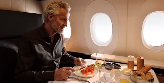 Mann mittleren Alters isst in einer First Class Suite mit Blick auf drei Fensterluken, vor ihm ein gedeckter Tisch mit Lachs-Vorspeise, Weißwein und Brotkorb.