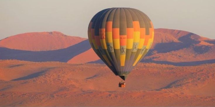 Heißluftballon schwebt über den roten Dünen einer Wüstenlandschaft bei Sonnenaufgang.