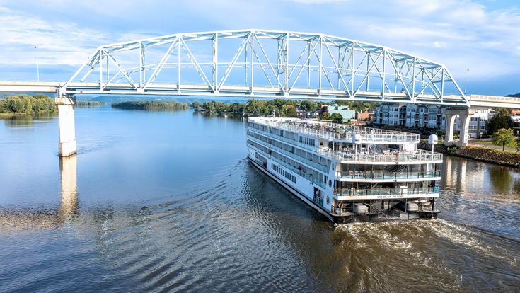 Flusskreuzfahrtschiff unter einer Brücke auf einem breiten, ruhigen Fluss – Symbol für gemächliches Reisen, Komfort und das Entdecken landschaftlicher Vielfalt entlang der Wasserwege.
