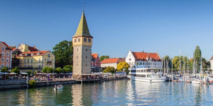 Blick auf den Hafen von Lindau am Bodensee mit Mangturm, Segelbooten und historischen Gebäuden bei blauem Himmel.