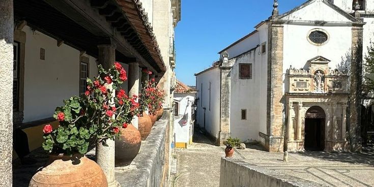 Historische Kirche an einer gepflasterten Gasse, gesäumt von alten Gebäuden und großen Tontöpfen mit roten Blumen.