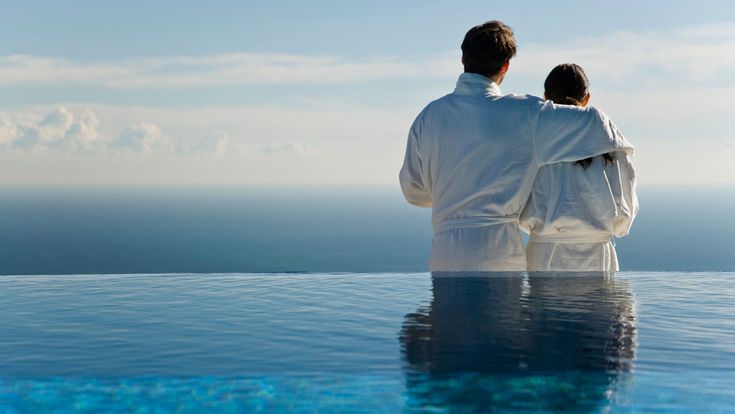 Paar im Bademantel blickt aufs Meer, davor Infinity Pool