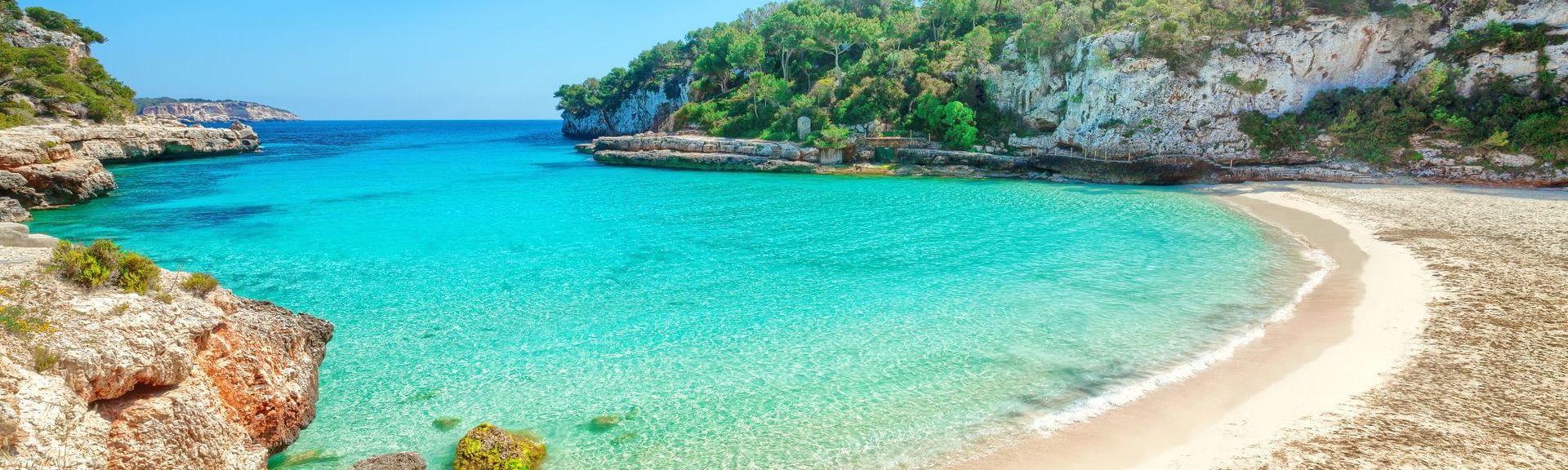 Eine idyllische Bucht auf Mallorca mit kristallklarem, türkisfarbenem Wasser, einem kleinen Sandstrand und umgeben von felsigen Klippen sowie grüner Vegetation unter strahlendem Sonnenschein.