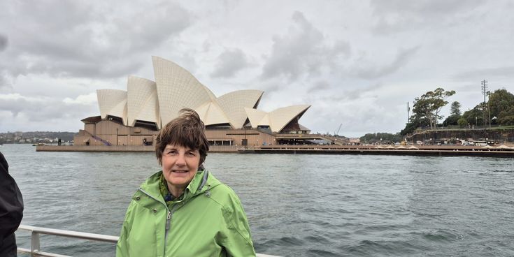 Petra Meyers steht auf einer Fähre mit Blick auf das Sydney Opera House in Australien, unter grauem Himmel über dem Hafen.
