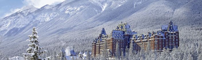 Verschneites Luxus-Berghotel (Fairmont-Stil) liegt in einem Wintertal vor hohen, weißen Gipfeln.