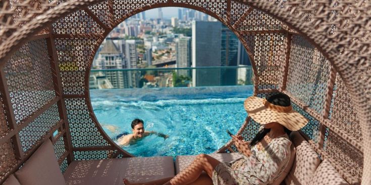 Frau mit Sonnenhut entspannt in einer geflochtenen Lounge am Dachpool, während ein Mann im Pool schwimmt.