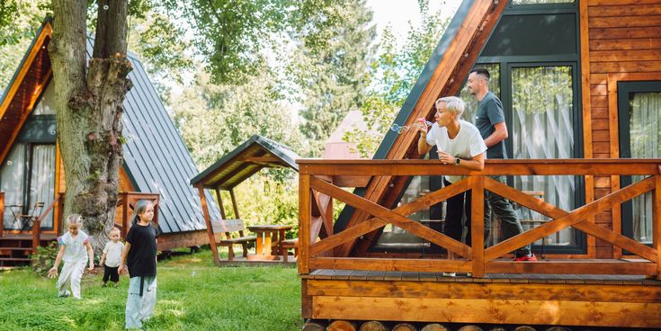 Familie vor Holzferienhäusern in der Natur – Erwachsene auf der Veranda, Kinder spielen lachend im Gras unter Bäumen.