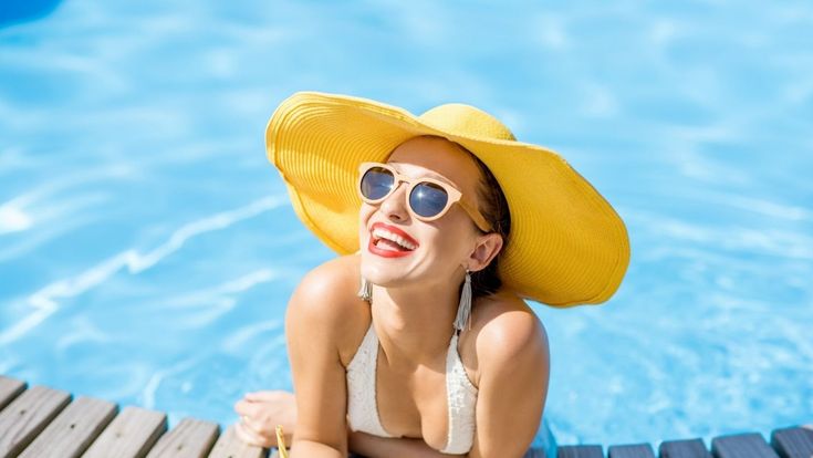 Lachende Frau mit gelbem Sonnenhut und Sonnenbrille entspannt am Rand eines blauen Pools.