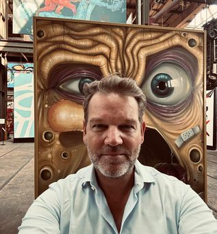 Ein Mann posiert vor einem farbenfrohen Kunstwerk mit einem Auge im Hintergrund.