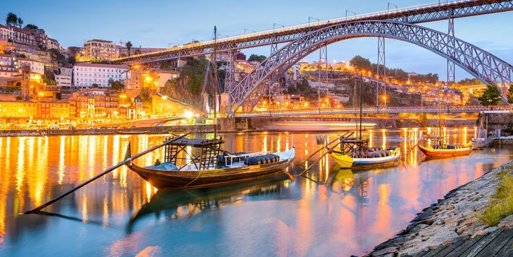 Traditionelle Rabelo-Boote auf dem Douro vor der beleuchteten Ponte Dom Luís I und der Altstadt von Porto in der Abenddämmerung.