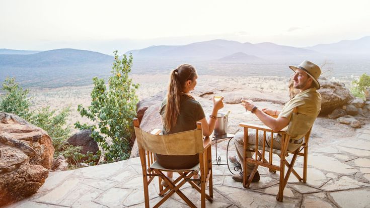Paar sitzt auf einer Terrasse mit weitem Blick über die afrikanische Savanne, genießt bei Sonnenuntergang ein Getränk und den Moment der Ruhe.