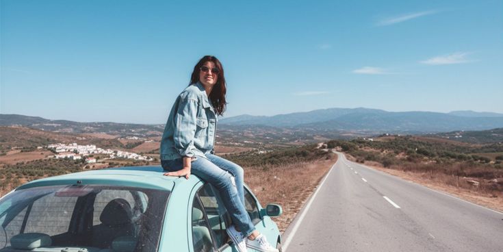 Frau mit Sonnenbrille sitzt auf einem türkisfarbenen Auto an einer einsamen Landstraße vor einer hügeligen Landschaft.
