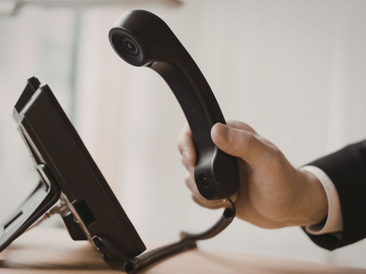 Hand nimmt den Hörer eines schwarzen Tischtelefons ab, das auf einem Schreibtisch steht – Nahaufnahme mit Fokus auf dem Telefonhörer und der Hand.