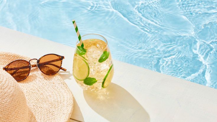 Sommerliche Szene am Pool mit Strohhut, Sonnenbrille und erfrischendem Getränk mit Limette und Minze auf einem hellen Tisch.